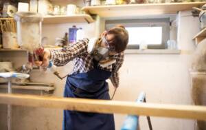 Young woman craftswoman applying varnish on wooden elements in woodshop