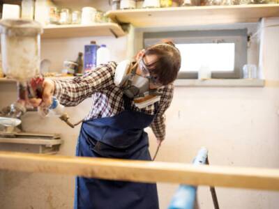 Young woman craftswoman applying varnish on wooden elements in woodshop